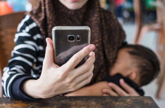 Mother wearing hijab using mobile phone while breastfeeding baby.